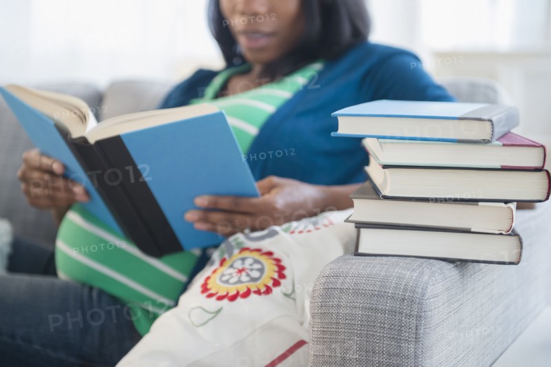 Black pregnant woman reading books on sofa