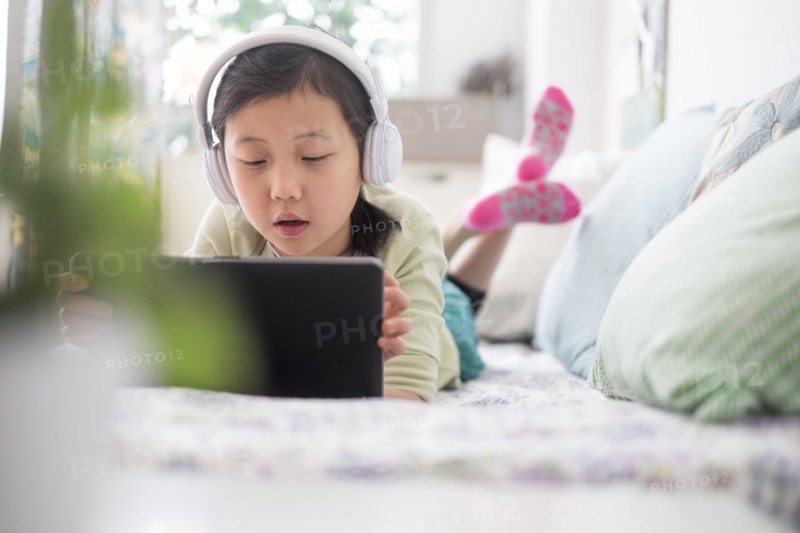 Chinese girl using digital tablet with headphones on bed