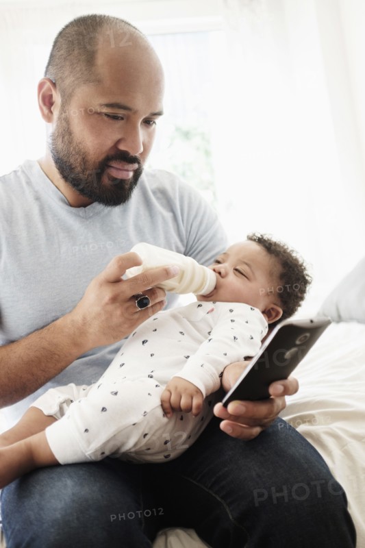 Father bottle feeding baby son and using cell phone