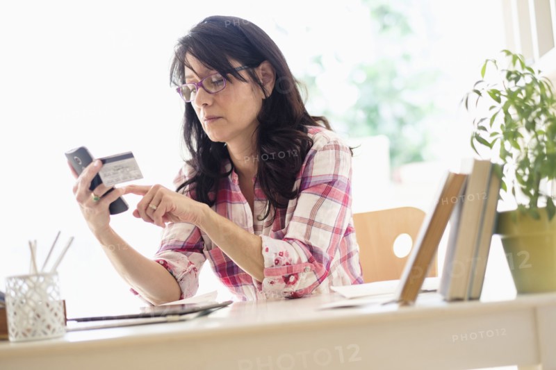 Hispanic woman using credit card with cell phone in home office