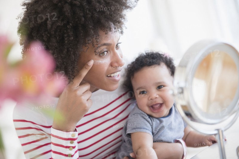 Mother holding baby son applying face cream in mirror