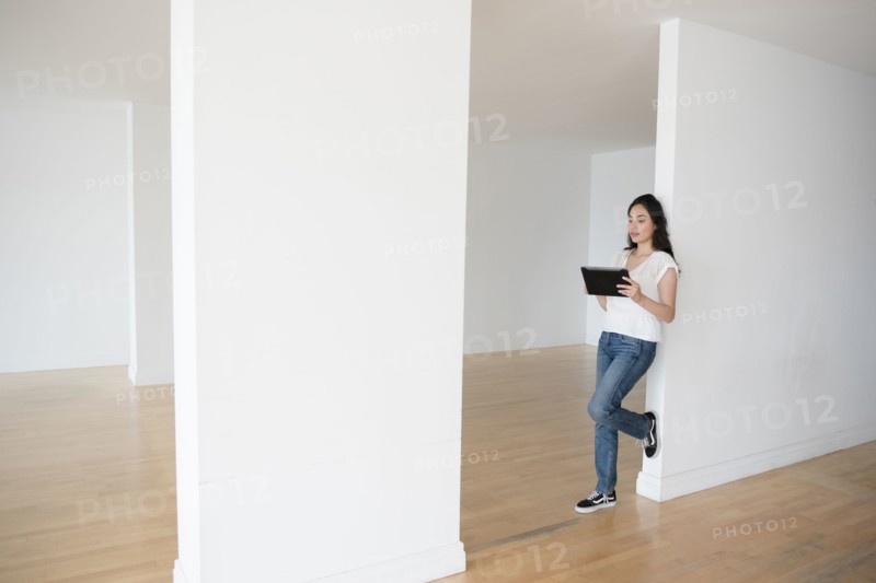 Hispanic woman leaning on wall of empty apartment holding digital tablet