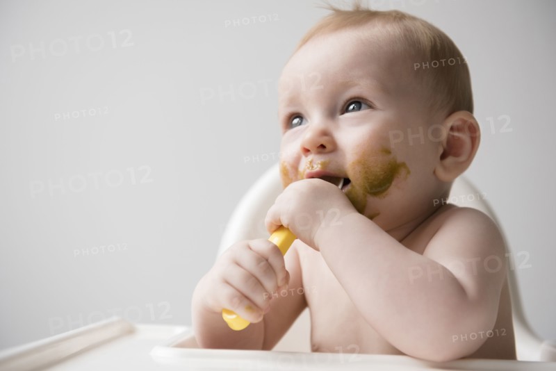 Messy Caucasian baby boy eating from spoon in high chair