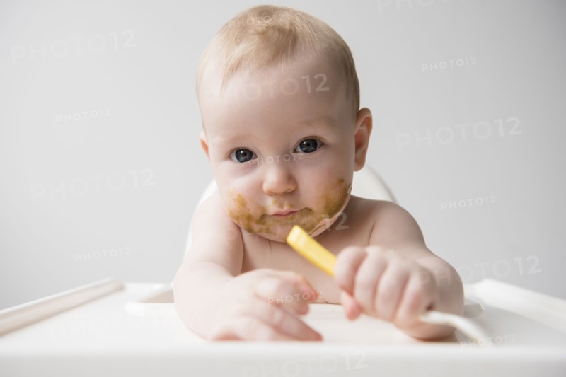 Messy Caucasian baby boy holding spoon in high chair