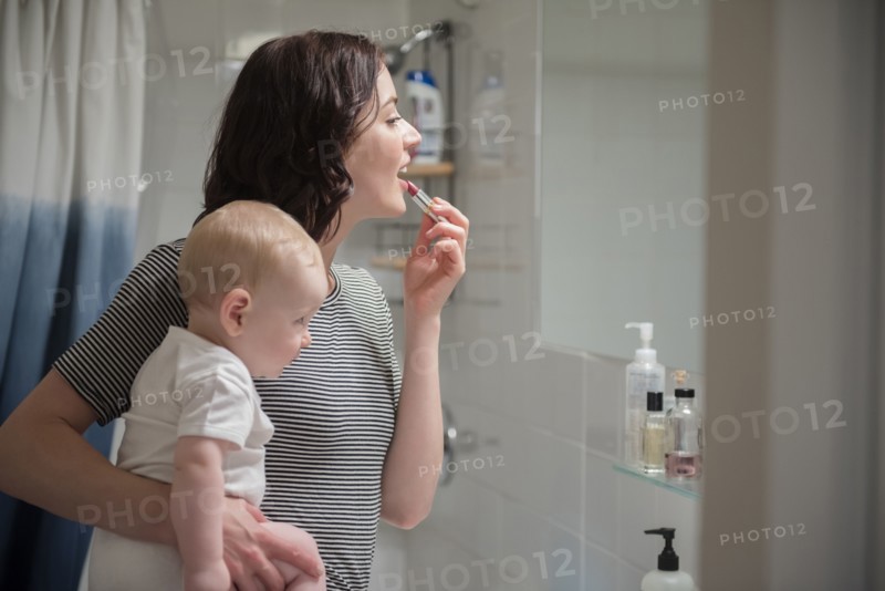 Caucasian mother holding baby son while applying lipstick in mirror