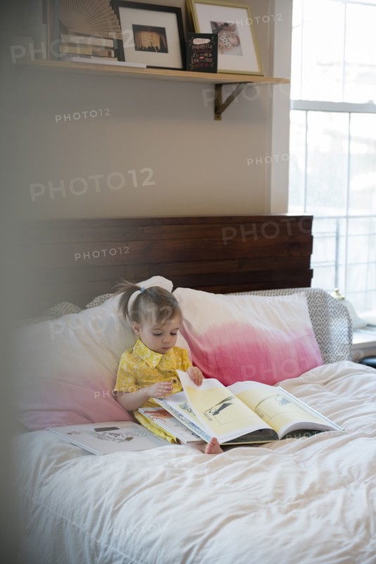 Caucasian baby girl sitting on bed reading book