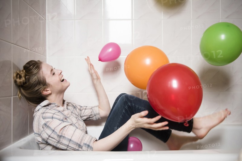 Laughing woman sitting in bathtub playing with multicolor balloons