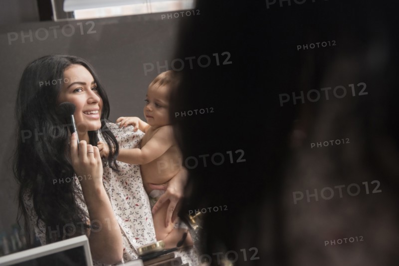 Caucasian mother holding baby son and applying makeup in mirror