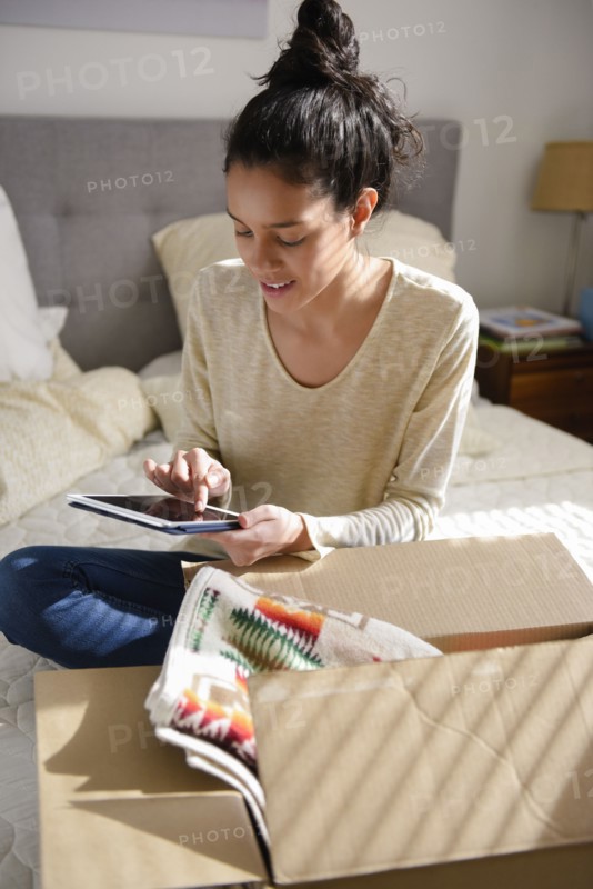 Hispanic woman sitting on bed near box using digital tablet