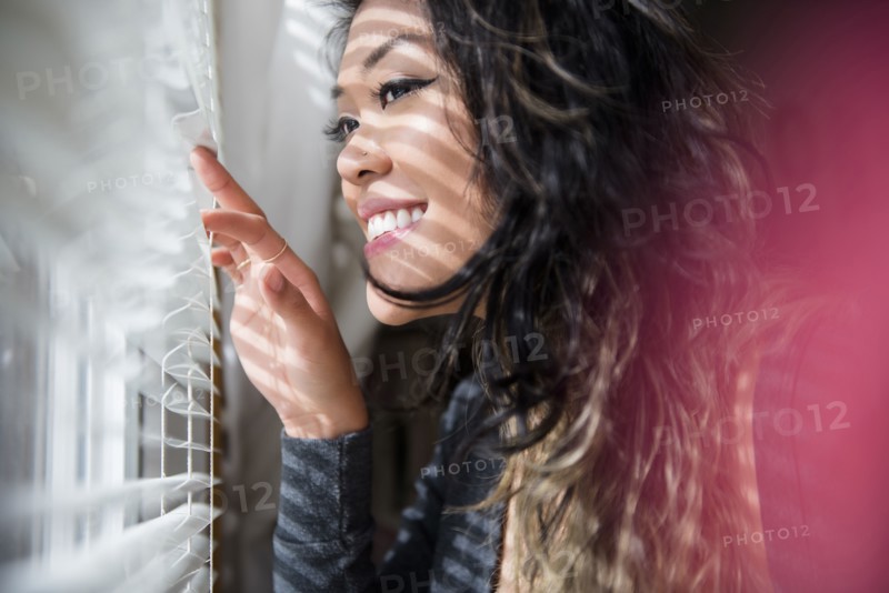 Mixed Race woman peeking out window behind blinds