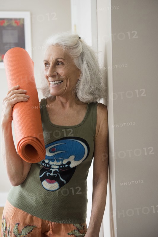 Older woman leaning on wall holding exercise mat