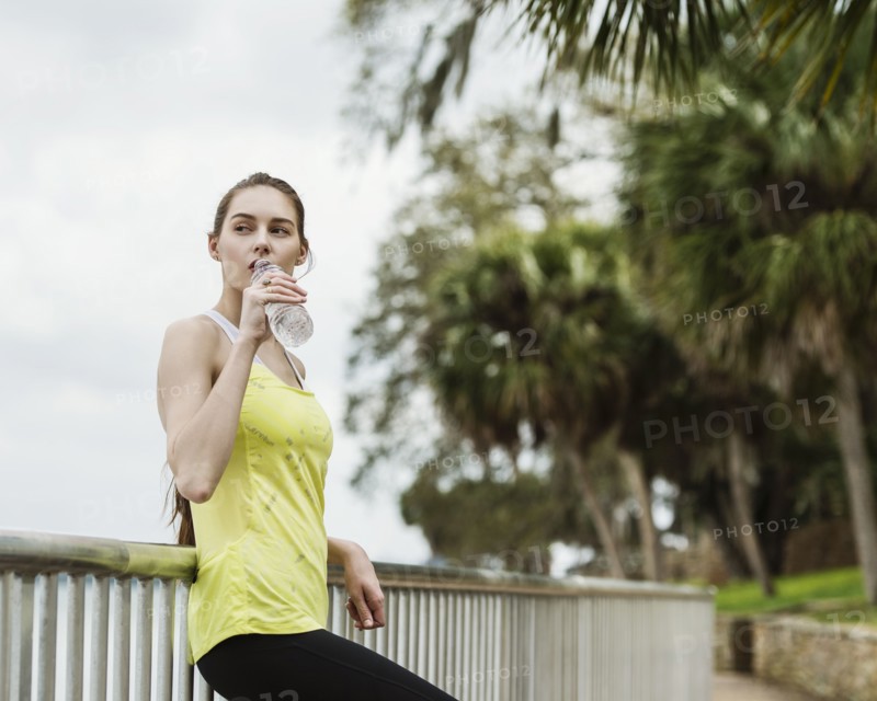 Woman in sports clothing leaning on fence and drinking water from bottle