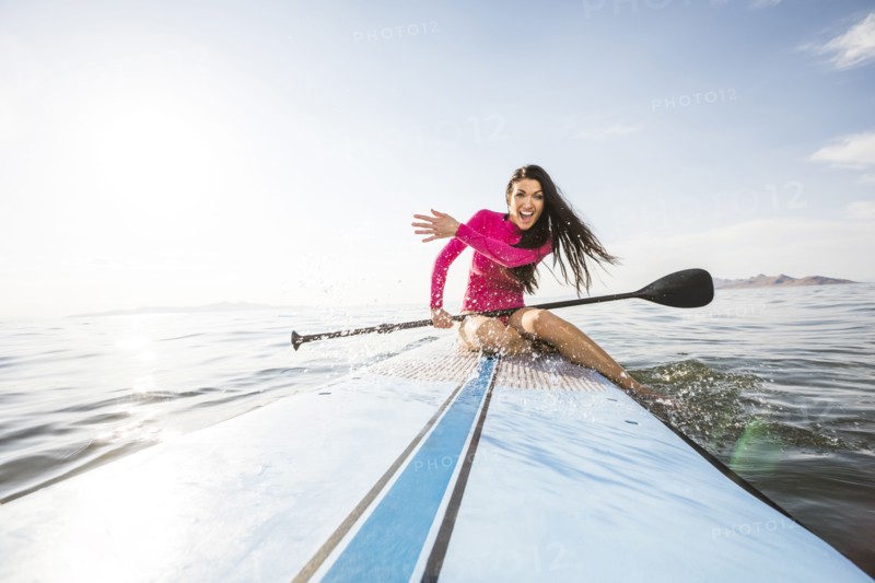 Smiling woman in pink swimsuit sitting on paddleboard and splashing water