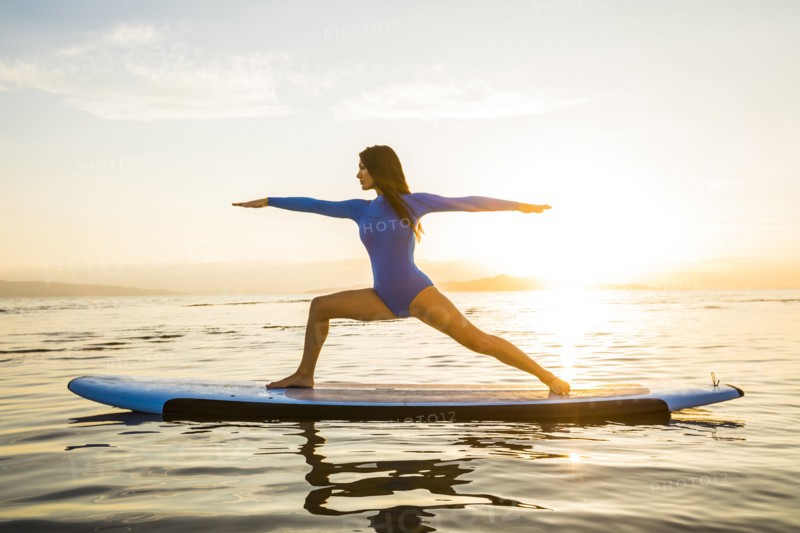 Woman in blue swimsuit performing Virabhadrasana II (Warrior II Pose) on paddleboard at sunset