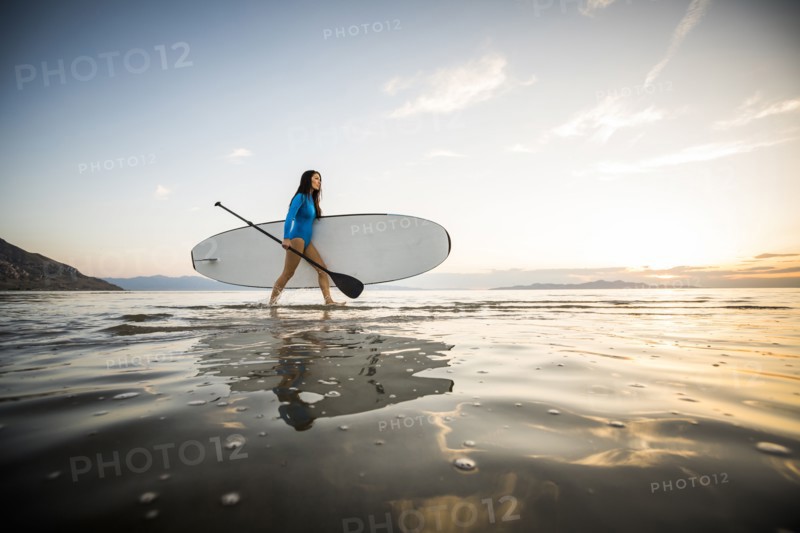 Woman in blue swimsuit carrying paddleboard in lake at sunset