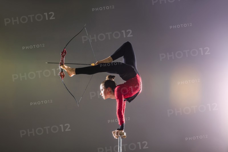 Young acrobat performing with archery bow on gymnastics bar