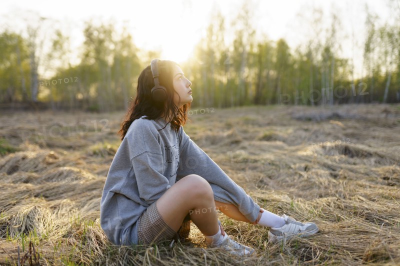 Side view of woman listening to music while sitting on grass in meadow at sunset
