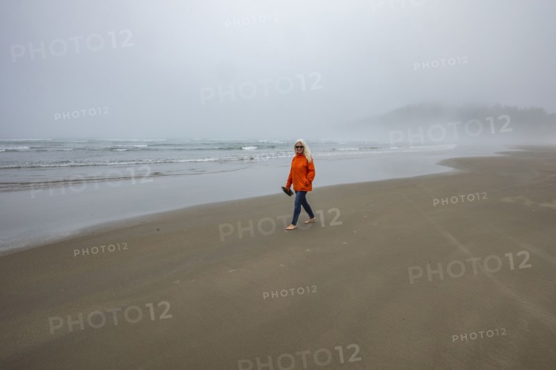 Woman in orange jacket walks along foggy beach