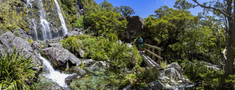 Rear view of hiker crossing footbridge near waterfall in Fiordland National Park, Te Anau, Fiordland, New Zealand