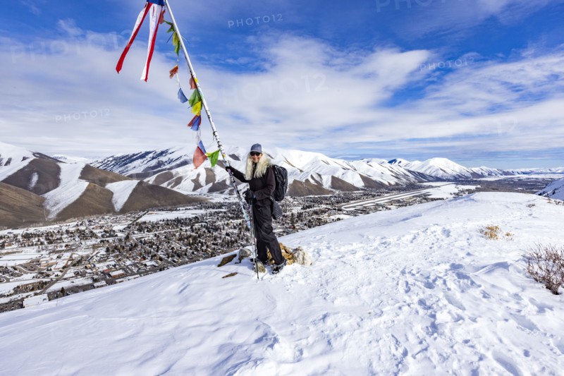 Smiling woman with snowshoes standing at pole with colorful flags on top of mountain, Hailey, Idaho, USA