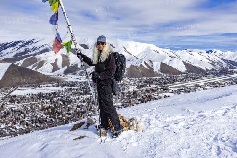 Smiling woman with snowshoes standing at pole with colorful flags on top of mountain, Hailey, Idaho, USA