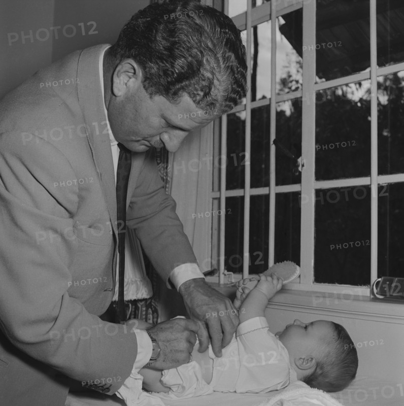 Changing Tubby Block's baby. Tubby Block's baby plays with a hairbrush whilst a man in a jacket and tie, possibly Tubby Block himself, changes it's nappy. Kenya, 30 August 1954. Kenya, Eastern Africa, Africa.