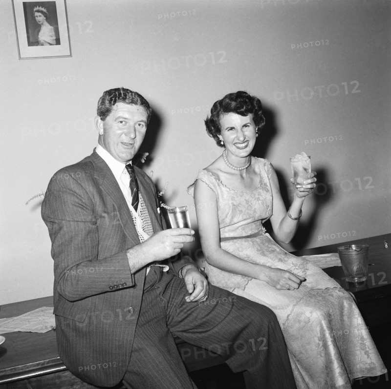 Cheers. Cheerful couple shown seated with drinks below a picture of Queen Elizabeth II at an RAF party buffet. Kenya, 27 November 1954. Kenya, Eastern Africa, Africa.