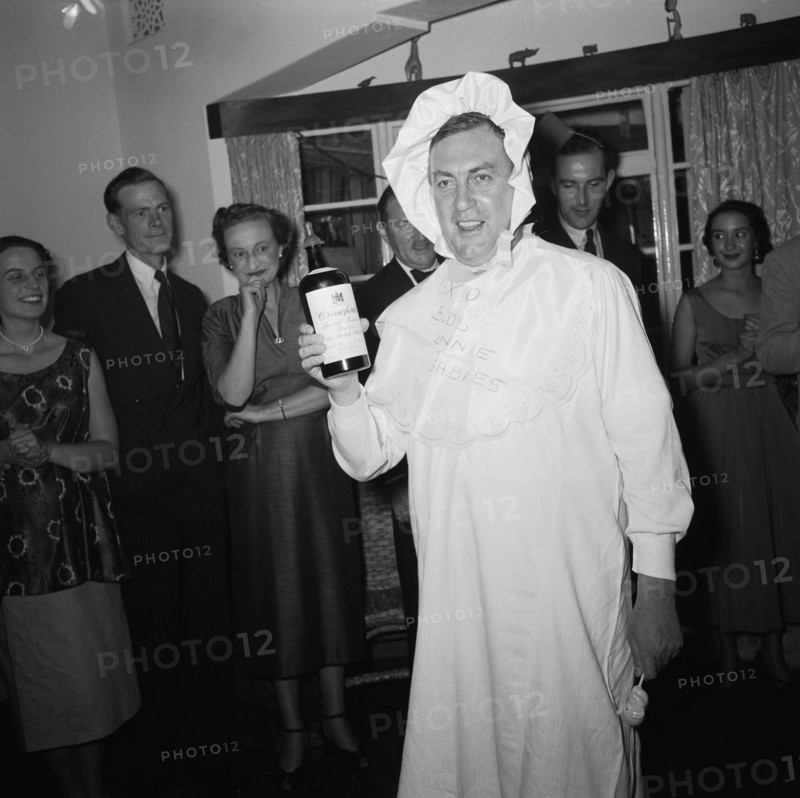 Baby costume. A man wears a fancy dress baby costume at the Archer's New Year party. He holds a bottle of Crawfords old scotch whiskey in one hand and an elephant-shaped baby's rattle in the other. A small crowd of onlookers in the background are clearly entertained. Kenya, 31 December 1954. Kenya, Eastern Africa, Africa.