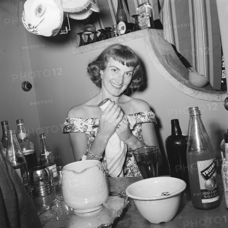 Drying up. A cheerful woman dries a glass at the Archer's New Year party. The table in front of her is covered with bottles and glasses and a selection of fancy dress animal masks hang over her head. Kenya, 31 December 1954. Kenya, Eastern Africa, Africa.
