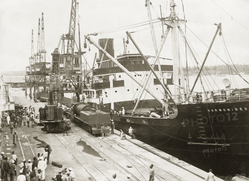 A train unloaded from SS Harmonides. A locomotive boiler and tender have been lowered onto a bogie on the dockside railway at Kilindini harbour by cranes onboard SS Harmonides. A railway steam crane stands by the train. Mombasa, Kenya, circa 1926. Mombasa, Coast, Kenya, Eastern Africa, Africa.