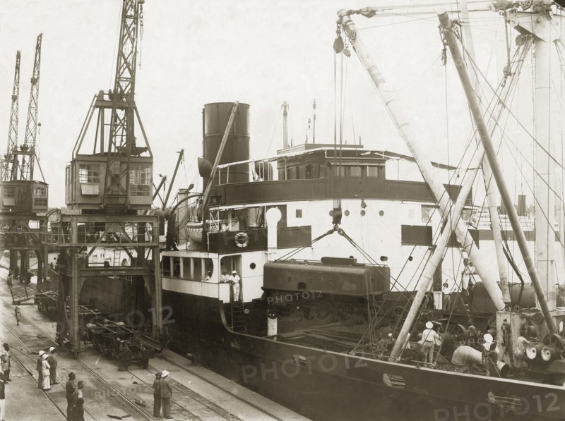 Unloading a locomotive tender from SS Harmonides. Cranes onboard the SS Harmonides hoist a locomotive tender (water tank railcar or wagon) above the ship's decks at Kilindini harbour. Mombasa, Kenya, circa 1926. Mombasa, Coast, Kenya, Eastern Africa, Africa.