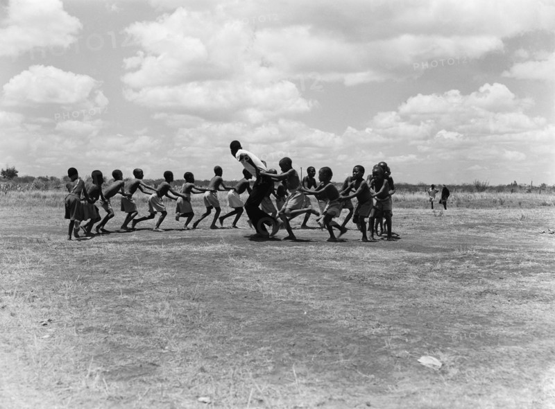 A train of children. A train of children wearing shorts hold onto one another as they run around in a circle, led by an adult. An original caption suggests this is a physical training lesson. Kenya, 25 February 1953. Kenya, Eastern Africa, Africa.