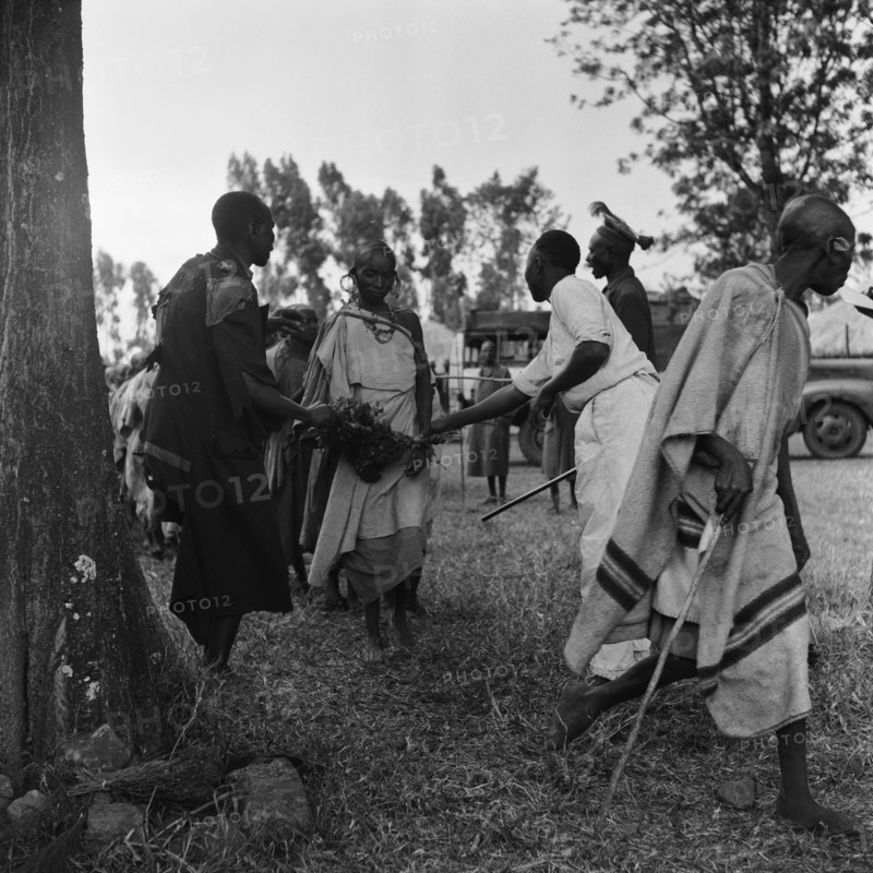 Cleansing ceremony. Two men brush bundles of branches against a woman wearing jewellery as she passes between them at a cleansing ceremony. Another man holding a baton shouts at a line of people queuing for the same treatment. Kenya, 20-28 March 1953. Kenya, Eastern Africa, Africa.
