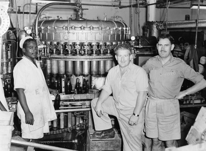 EA Breweries. Three men stand next to brewing machinery at an EA Breweries factory. The black worker pictured left holds a bottle and wears an apron printed with the letters 'EA'. Kenya, 21 April 1953. Kenya, Eastern Africa, Africa.