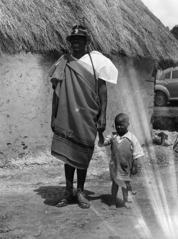 Chief Makimei. Chief Makimei holds the hand of a young boy. He wears a blanket wrapped around one shoulder and carries a shotgun on a strap over his arm. A metal badge in the shape of a lion is pinned to his hat and reads 'CHIEF'. Kenya, 12 May 1953. Kenya, Eastern Africa, Africa.