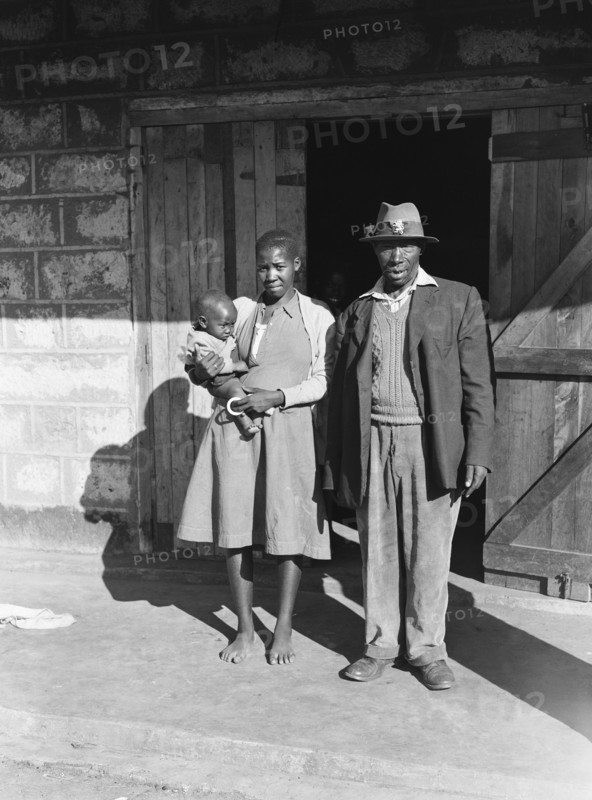 Headman Paulo and wife. Headman Paulo pictured outside the doorway of a building with his barefoot wife who holds a baby. Kenya, 17 May 1953. Kenya, Eastern Africa, Africa.