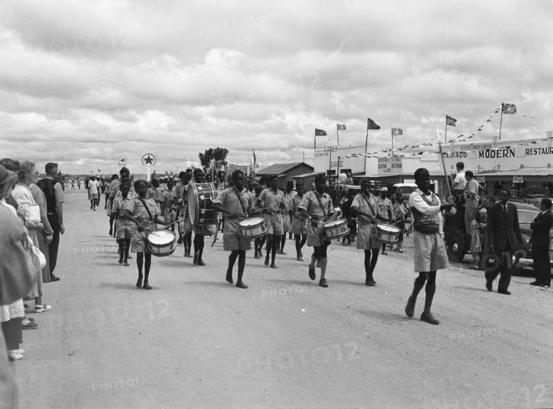 Maasai ngoma. Onlookers at a traditional 'ngoma' watch as a group of young Maasai people wearing uniforms pass through the centre of town playing drums. Kajiado, Kenya, 2 June 1953. Kajiado, Rift Valley, Kenya, Eastern Africa, Africa.