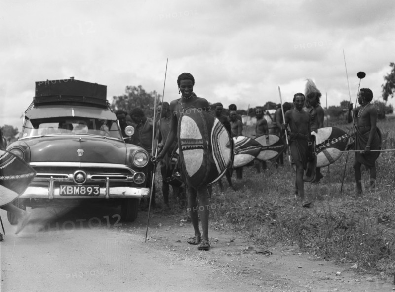 Maasai and Ford. Maasai warriors holding spears and shields congregate on the roadside next to a Ford car at a 'ngoma', a word meaning drum that is also used to describe traditional celebrations. Kajiado, Kenya, 2 June 1953. Kajiado, Rift Valley, Kenya, Eastern Africa, Africa.