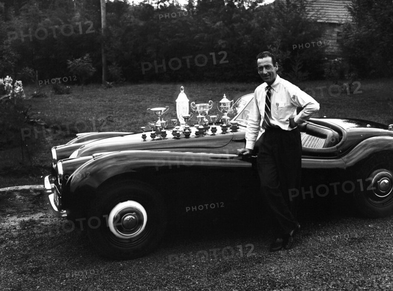 Manussis with trophies. John Manussis leans on the wing of a Jaguar Coupe convertible car, the bonnet of which displays numerous trophies. Kenya, 7 June 1953. Kenya, Eastern Africa, Africa.