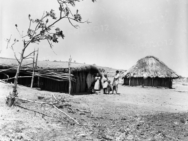Mbulu hut. An adult and three children stand outside a long wooden hut with a flat, thatched roof. Kenya, August 1953. Mbulu, Rift Valley, Kenya, Eastern Africa, Africa.