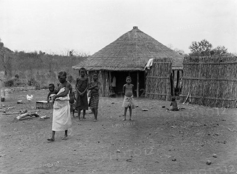 Mozambique natives. A group of children outside a wooden hut with a thatched roof. A barefooted woman in the foreground carries a baby on her hip as she walks. Mozambique, September 1953. Mozambique, Southern Africa, Africa.