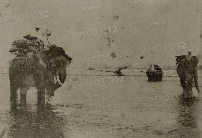 Hunting elephants wet their feet. Two hunting elephants, laden with blankets, cool their feet in a shallow river, their mahouts (elephant handlers) sitting patiently on their shoulders. North East India, circa 1890. India, Southern Asia, Asia.