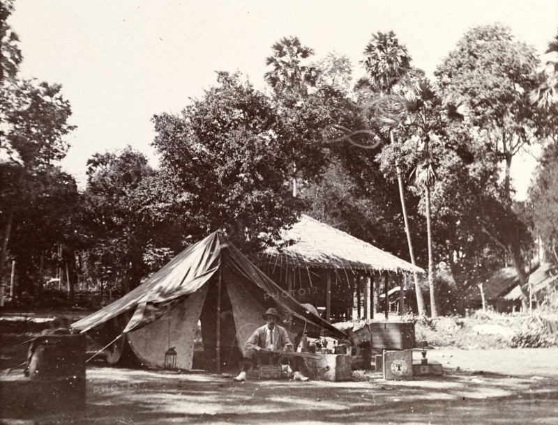 Camping in a village. A European man sits on an upturned crate outside his tent, brandishing a shotgun. The thatched roofs of dwellings in the distance suggest he may be camping in a rural village. Possibly Burma (Myanmar), circa 1908., South East Asia, Asia.
