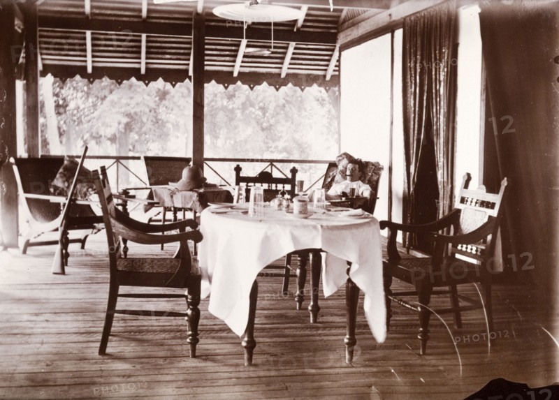 Colonial veranda. A European man smokes a cigar as he reclines in a chair on a colonial veranda. His shotgun rests upright against an armchair: a dining table set for two dominates the foreground. Possibly Burma (Myanmar), South East Asia, circa 1910., South East Asia, Asia.