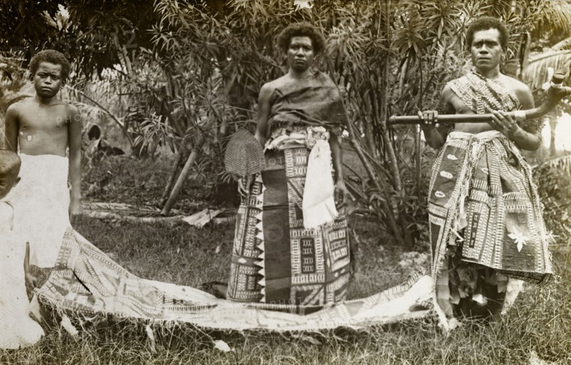 Fijian ceremonial costume. A Fijian man and woman pose for the camera dressed in traditional ceremonial attire. The woman holds up a basketwork fan, the man a curved stick. Fiji, circa 1920. Fiji, Pacific Ocean, Oceania.