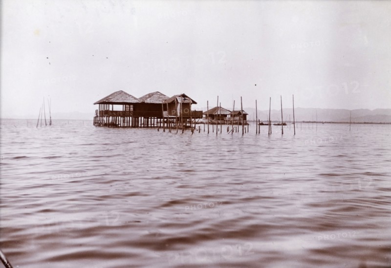 Lake dwellings in Burma (Myanmar). Two wooden dwellings on stilts project from the calm waters of a lake. Burma (Myanmar), circa 1910. Mohnyin, Kachin, Burma (Myanmar), South East Asia, Asia.
