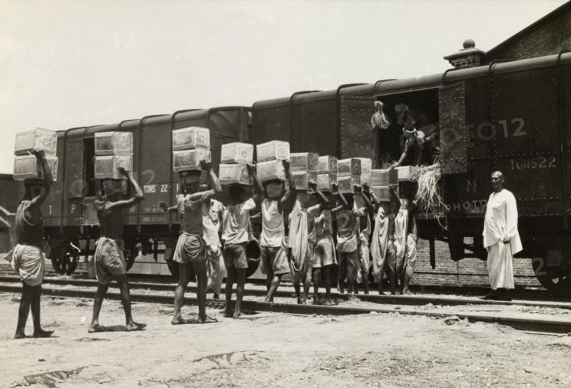 Queuing up to load a freight train. A team of workers carry boxes on their heads and form a queue, as they load cargo onto a freight car under the direction of an overseer. India, circa 1942. India, Southern Asia, Asia.