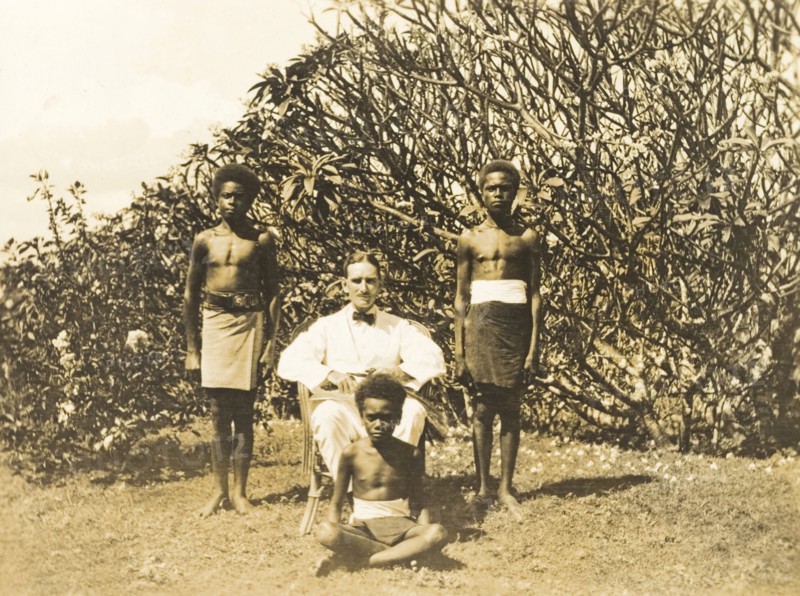Clifford Francis poses with his servants. Outdoors portrait of Clifford Francis, a European District Officer, posing in a chair beside three of his Solomon Islander domestic servants. Gizo, Solomon Islands, 1919. Gizo, West (Soloman Islands), Solomon Islands, Pacific Ocean, Oceania.