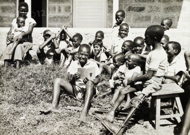 Children at a charity in Nairobi. An older boy reads a story to a group of younger children on a patch of grass outdoors. A disabled boy to his left sits on a stool with his leg bound in a splint. An original caption comments that this boy 'had appealed to us to be able to wear (his crutches) and so walk again', after his father had pawned his walking aids twice, and suggests that these children were receiving help from a European charity of some sort. Nairobi, Kenya, March 1955. Nairobi, Nairobi Area, Kenya, Eastern Africa, Africa.