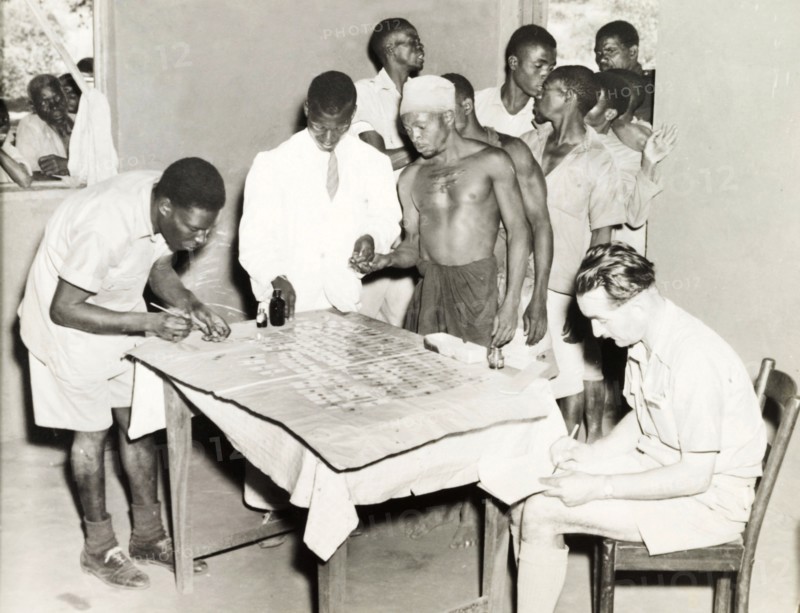 Blood samples at village dispensary. A group of African men queue at a dispensary, where two African medics are taking blood samples. A European doctor sits writing notes next to a table, on which the blood samples are laid out on slides. Probably Northern Rhodesia (Zambia), circa 1950. Zambia, Southern Africa, Africa.